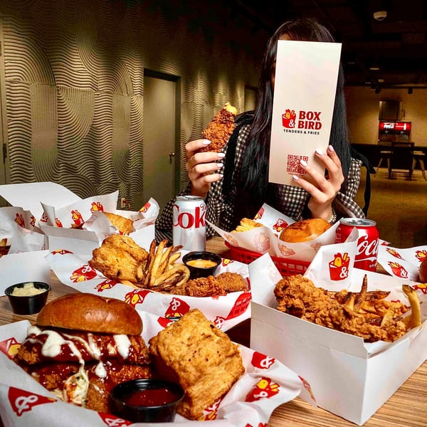 A person holds a Box & Bird menu, surrounded by fried chicken, fries, and sandwiches on a table.