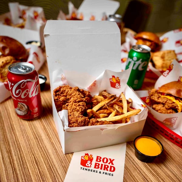 Box & Bird fried chicken tenders and fries in a box, with drinks and burgers on a wooden table.