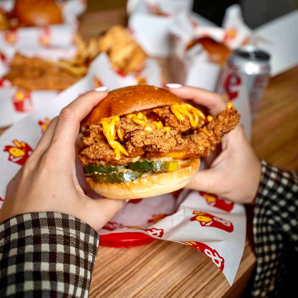 Hands holding a Box & Bird fried chicken sandwich with pickles and cheese sauce.